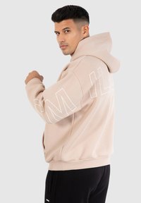 Sweat à capuche beige avec des manches longues, grand lettrage blanc le long de la manche, et bords côtelés au niveau des poignets et de l'ourlet. Porté avec un pantalon noir.