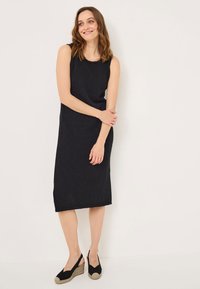 Černé bezrukávové šaty s rovnou siluetou, texturovaný pletený materiál a lem končící nad kotníky. Nosené s černými espadrilkami.