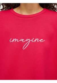 Rosa genser med hvit brodert tekst som sier «imagine». Stoffet er glatt, med rund hals og korte ermer.