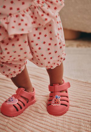 Roze, flexibele sandalen met een open ontwerp en twee verstelbare riemen. Voorzien van een hart- en lieveheersbeestjeversiering aan de zijkant. Geprofileerde zool.