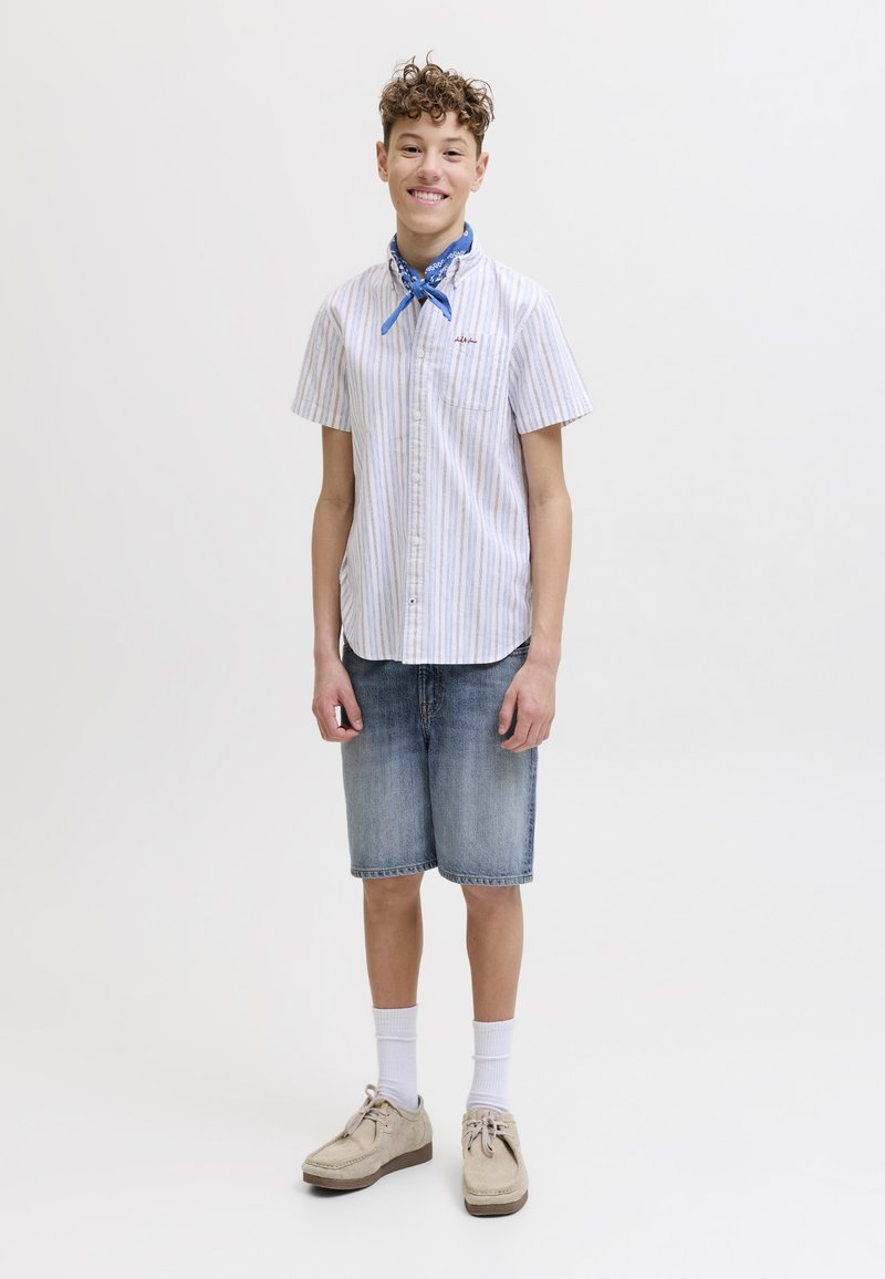 Garçon adolescent aux cheveux bouclés souriant, portant une chemise à manches courtes rayée, un bandana bleu, un short en jean, des chaussettes blanches et des chaussures beiges.