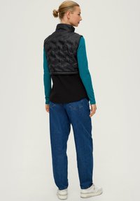 Schwarz gesteppte Weste mit hohem Kragen, kombiniert mit einem tealfarbenen Langarmshirt und lockeren blauen Jeans, die eine bequeme Passform und ein einfaches Design aufweisen.