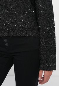 Maglione nero scintillante con dettagli in filo metallico e polsini a coste, abbinato a jeans neri con chiusura a due bottoni. Superficie texture.