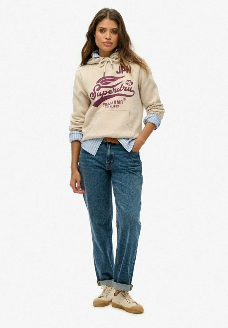 Beige hoodie met logo, koordjes en voorzak; gelaagd over een blauw gestreept shirt. Draagt met lichtblauwe jeans en beige sneakers.
