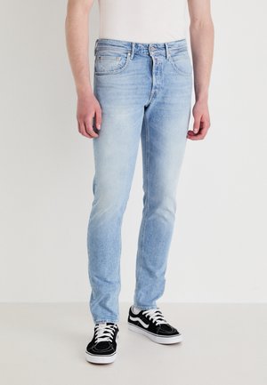 Jasnoniebieskie dżinsy o slim fit, wyposażone w pięć kieszeni, z subtelnym przetarciem, zestawione z czarnymi sneakersami z białymi akcentami.