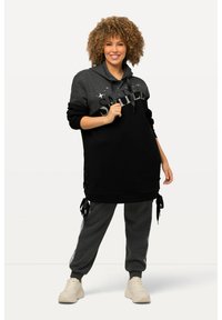 Schwarzes und graues Kapuzenkleid mit dem Aufdruck "SMILE", Sternelementen, seitlichen Schnürungen, kombiniert mit grauen Jogginghosen und hellen Sneakern.