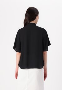 Blusa nera realizzata in tessuto leggero, con maniche corte a sbuffo e colletto alto. La vista posteriore mostra una vestibilità ampia con dettagli minimi.