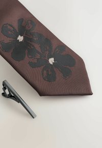 Brown tie featuring a black floral pattern, made of textured fabric. Accompanied by a silver metal tie clip with a sleek design.