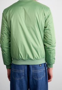 Giacca bomber verde in un tessuto liscio e lucido; polsini e orlo elasticizzati; presenta una sottile trama, abbinata a jeans blu dal taglio comodo.