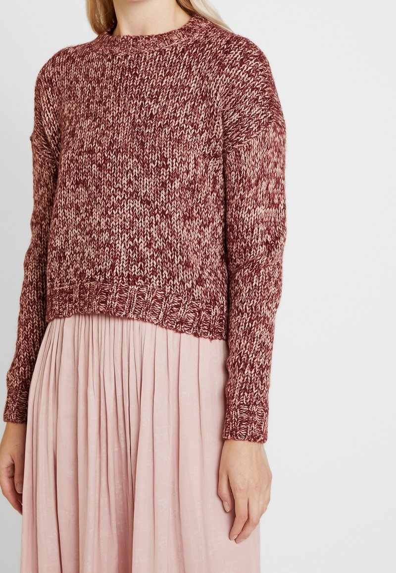 Donna che indossa un maglione a maniche lunghe a maglia mélange bordeaux e rosa, abbinato a una gonna plissettata rosa chiaro, in piedi davanti a uno sfondo bianco.