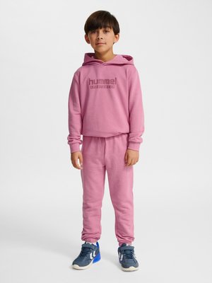 Garçon debout portant un sweat à capuche hummel rose et un pantalon de survêtement assorti avec des baskets bleu marine sur fond blanc.
