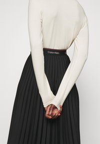 Top a maniche lunghe crema abbinato a una gonna plissettata nera con una vita decorata dal marchio Calvin Klein in bianco e rosso.