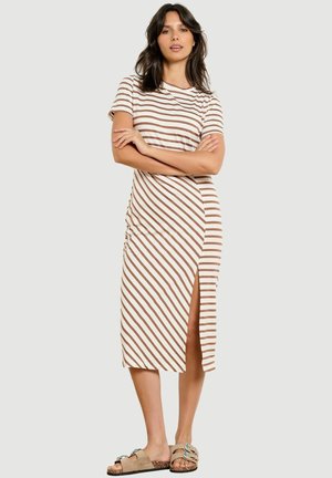 Žena v béžovom a bielom pruhovanom midi šatách so štrbinou na boku, so založenými rukami, obutá v béžové otvorené sandále na svetlom jednofarebnom pozadí.