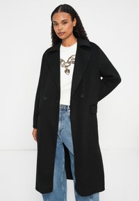 Mujer vestida con un abrigo largo negro sobre un suéter blanco con un estampado de cadenas doradas y jeans azules, de pie contra un fondo blanco.