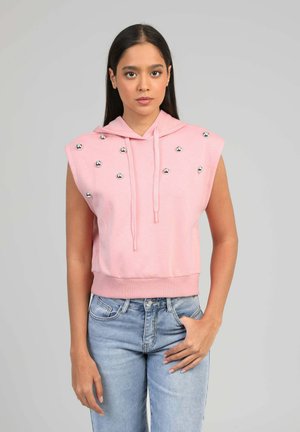 Femme aux longs cheveux foncés portant un sweat à capuche rose sans manches avec des clous argentés et un jean bleu, debout devant un fond gris uni.