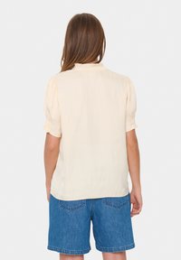 Crème blouse met korte mouwen, een gerimpelde kraag en elastische manchetten, gecombineerd met blauwe denim shorts. Gladde textuur, eenvoudig ontwerp.