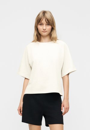 Ung kvinde med skulderlangt blondt hår iført en off-white kortærmet top og sorte shorts, stående foran en ensfarvet baggrund.