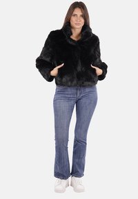Giacca cropped in eco-pelliccia nera con colletto ampio, abbinata a jeans blu e sneaker bianche. Presenta maniche larghe e una superficie texturizzata.