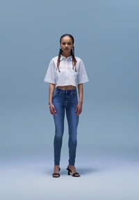 Camisa branca curta com botões, jeans skinny azul e sandálias pretas de salto; modelo está de pé contra um fundo azul claro.