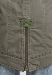 Gros plan sur la fermeture éclair d'une veste vert olive avec un patch rectangulaire cousu au-dessus, portée sur un jean en denim bleu clair.