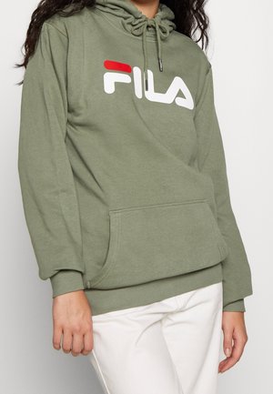 Osoba nosící olivově zelenou mikinu FILA s kapucí a přední kapsou a bílé kalhoty, stojící proti jednobarevnému světlému pozadí.