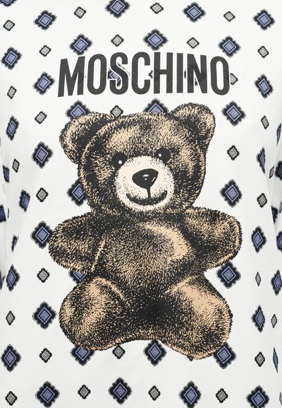 Λευκό T-shirt με μεγάλο καφέ λούτρινο αρκουδάκι και το κείμενο "Moschino", περιβαλλόμενο από μοτίβα σε σχήμα διαμαντιού μπλε και μαύρου χρώματος.