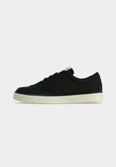 Μαύρο sneaker χαμηλού ύψους με μαύρα κορδόνια και παχύ ελαστικό πέλμα σε εκρού χρώμα, εμφανίζεται από το πλάι πάνω σε απλό ανοιχτό φόντο.