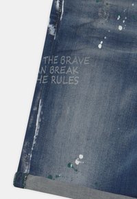 Sininen denim-kangas, jossa on valkoisia maalitippoja, kuluneita reunoja ja haalistunut teksti, jossa lukee "ROHKEAT VOIVAT RIKKOA SÄÄNTÖJÄ."