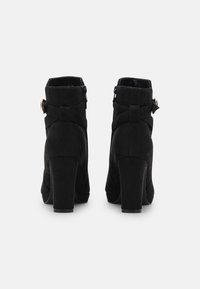 Botines de ante negro con tacón alto de bloque, cremalleras dobles en los laterales y detalles decorativos de correas en la parte trasera. Textura suave y diseño sencillo.