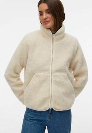 Kvinde iført cremefarvet fleecejakke med lynlås og forlommer samt blå jeans, stående med hænderne bag ryggen.