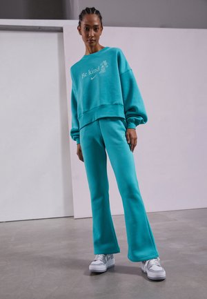 Conjunto de sweatshirt turquesa e calças flare em mistura de algodão. Apresenta o texto "Seja gentil" e o logo da Nike. Usado com ténis brancos. Textura suave.