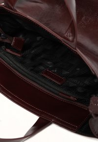 Tote en cuir bordeaux avec une finition brillante, présentant un intérieur noir à motif, une poche zippée et deux poignées solides.