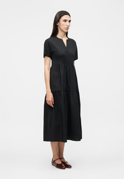 Γυναίκα με μακριά σκούρα μαλλιά, ντυμένη με μαύρο midi φόρεμα με κοντά μανίκια και σκούρα κόκκινα παπούτσια Mary Jane, στέκεται μπροστά σε απλό φωτεινό φόντο.