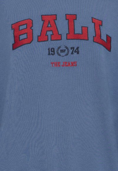 T-shirt en coton bleu avec un grand texte brodé en rouge « BALL », « EST 1974 » et « THE JEANS ». La texture est lisse avec une légère élasticité.