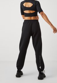 Žena v černém zkráceném topu s výřezem na zádech a černých joggingových kalhotách s logem "Stella McCartney" na noze, stojící na jednobarevném pozadí.