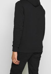 Sudadera negra con textura suave, que cuenta con un bolsillo canguro, puños y dobladillo acanalados. Diseñada para un ajuste relajado, sin marcas visibles.