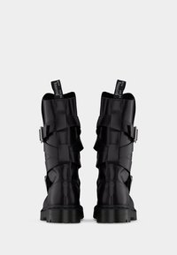 Ζευγάρι μαύρα δερμάτινα στρατιωτικά μποτάκια μεσαίου μήκους με ιμάντες με αγκράφες και ετικέτες τράβηξης με την ένδειξη "WITH SOLES Bouncing" σε απλό φόντο.