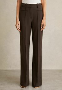 Pantaloni su misura marrone scuro a vita alta con pinces davanti, passanti per cintura, tasche laterali e gambe larghe, indossati con scarpe beige con punta appuntita.