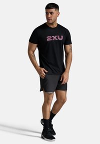 Svart kortärmad t-shirt med en texturerad rosa "2XU"-logotyp, tillsammans med mörka shorts och svarta träningsskor med vita accenter.