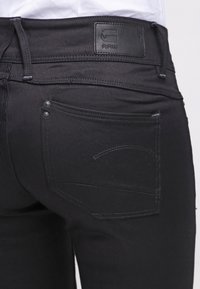 Los vaqueros de denim negro presentan una etiqueta de cuero con un logotipo en relieve en la parte trasera de la cintura, dos botones metálicos en el bolsillo trasero y detalles de costura sutiles.