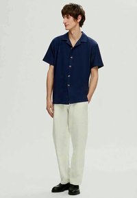 Camisa de manga curta azul-marinho com colarinho e cinco botões; usada com calças creme claras; modelo em pé com um fundo neutro.