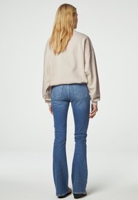 Felpa oversize beige con spalle abbassate, abbinata a jeans a zampa d'elefante blu con tasche posteriori, esposta su uno sfondo semplice.