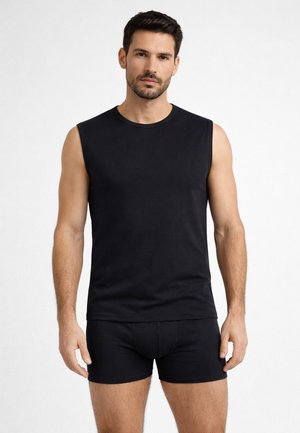 Uomo con canottiera nera e pantaloncini aderenti neri in piedi contro uno sfondo bianco uniforme.