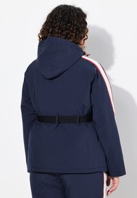 Námornická bunda s kapucí a páskem v pase, se světle modrými a červenými bočními pruhy, hladkou texturou a uvolněným střihem.