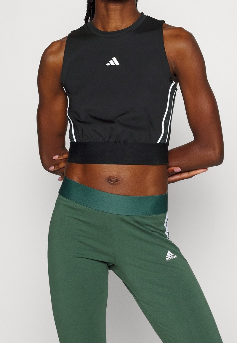 Personne athlétique portant un crop top noir sans manches et des leggings verts taille haute avec des logos et des rayures blanches, debout, les bras derrière le dos.