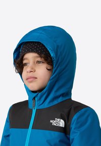 Modrá zateplená bunda s čiernym panelom, kapucňou a predným zipsom. Na hrudi je logo The North Face a pod bundou má textúrovanú pletenú čiapku.