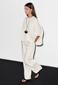 Une femme au style élégant marche avec confiance dans une tenue ample de couleur gris clair, ornée d'un collier en forme de cœur noir, le tout sur fond minimaliste.