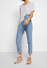 Mujer vestida con jeans ligeros azul claro arremangados, blusa de manga corta con lunares blancos y sandalias blancas de tacón bajo de tiras, de pie.