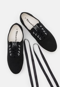 Svarta kanvas sneakers med vita gummisålar, rund tå och sex öglor för svarta snören. Varumärkeslogotyp synlig inuti. Snören ligger bredvid.