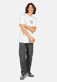 Weißes T-Shirt mit kreisförmigem schwarzem Grafikdesign, graue Cargo-Hosen mit mehreren Taschen und schwarz-weißen Sneakers.